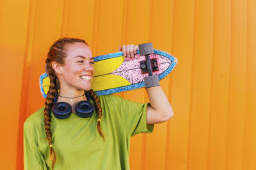 Fröhliche Frau mit Skateboard vor einer orangefarbenen Wand - MGRF00427