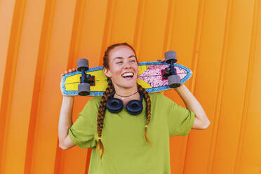 Junge Frau mit Skateboard vor einer orangefarbenen Wand - MGRF00428