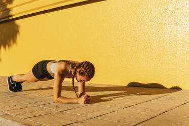 Sportswoman practicing plank on footpath during sunny day - MGRF00435