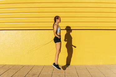 Young sportswoman using rope while skipping by yellow wall during sunny day - MGRF00441