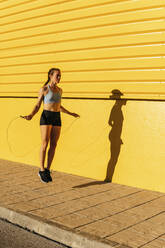 Female athlete using jump rope while practicing skipping by yellow wall - MGRF00442