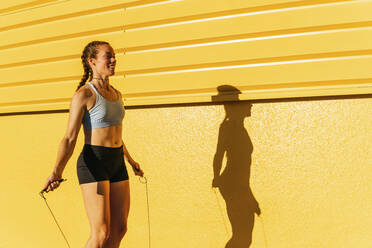 Smiling sportswoman jumping while using skipping rope by yellow wall - MGRF00444