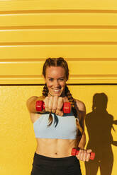 Glückliche Sportlerin, die mit einer Hantel vor einer gelben Wand trainiert - MGRF00446