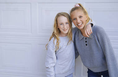Smiling girls with blond hair standing in front of white wall - AZF00368