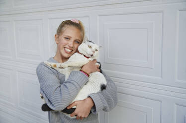 Blondes Mädchen, das eine Katze umarmt, während es an einer weißen Wand steht - AZF00371