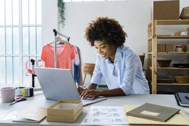 Junge Modedesignerin benutzt einen Laptop, während sie sich im Atelier auf den Schreibtisch stützt - GIOF13455