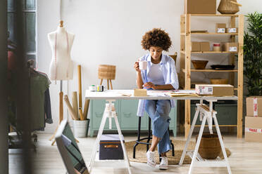 Modedesignerin mit Tasse am Schreibtisch im Atelier sitzend - GIOF13473