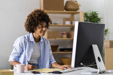 Junge Unternehmerin schreibt auf einem Briefumschlag, während sie einen Desktop-Computer im Studio benutzt - GIOF13482