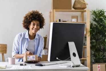 Businesswoman writing on package while looking at desktop computer in studio - GIOF13484