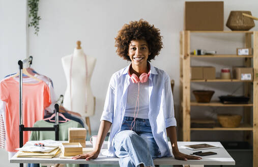 Happy young female entrepreneur with headphones sitting on desk at studio - GIOF13499