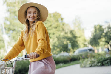 Smiling young woman wearing sun hat sitting on bicycle - VPIF04830