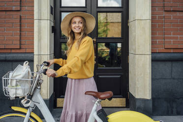 Beautiful woman looking away while standing with bicycle in front of door - VPIF04832