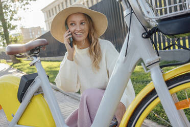 Lächelnde blonde Frau, die mit einem Smartphone spricht, während sie am Fahrrad hockt - VPIF04854