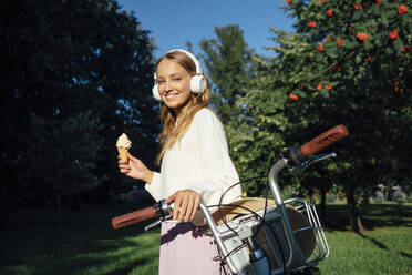 Young woman wearing headphones holding ice cream by bicycle at park during sunny day - VPIF04862