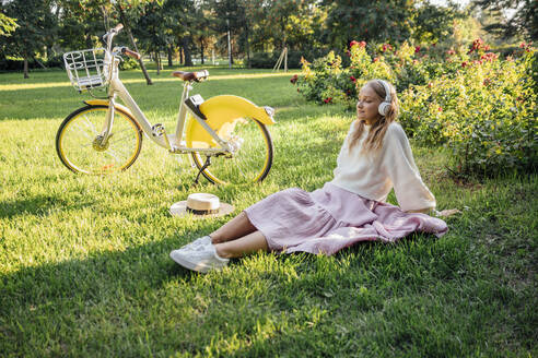 Junge Frau hört Musik über drahtlose Kopfhörer, während sie mit dem Fahrrad im Park sitzt - VPIF04864