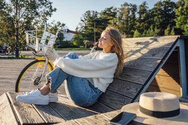 Young woman with eyes closed hugging knees while sitting on wooden bench - VPIF04886