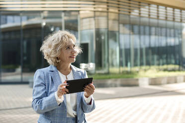 Lächelnde blonde Geschäftsfrau mit digitalem Tablet vor einem Bürogebäude - LLUF00066