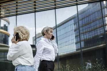 Businesswomen talking on mobile phones while walking by office building - LLUF00068