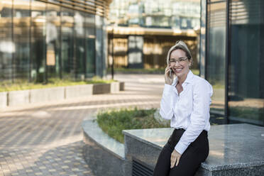 Lächelnde weibliche Fachkraft, die mit ihrem Smartphone vor einem Bürogebäude sitzt - LLUF00073
