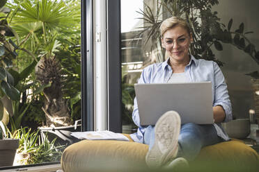 Lächelnde Freiberuflerin, die einen Laptop benutzt und am Fenster sitzt - UUF24694