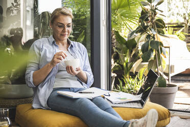 Geschäftsfrau trinkt Kaffee und sitzt mit Laptop und Tagebuch am Fenster - UUF24698