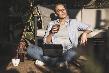 Thoughtful female freelancer holding drink with digital tablet on terrace - UUF24703