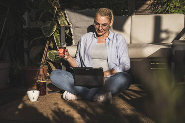 Ältere Geschäftsfrau hält ein Getränk in der Hand und sitzt mit einem digitalen Tablet auf einer Terrasse - UUF24704