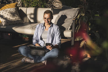 Lächelnde Geschäftsfrau, die auf einer Terrasse ein Mobiltelefon und ein digitales Tablet benutzt - UUF24705