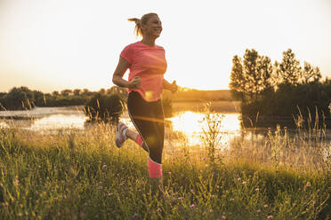 Lächelnde Frau joggt bei Sonnenuntergang auf einer Wiese - UUF24709