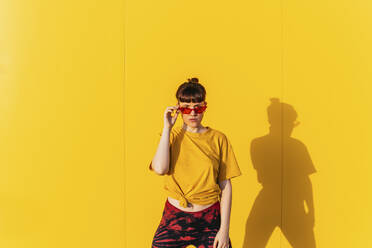 Mid adult woman wearing sunglasses in front of yellow wall during sunny day - MGRF00467