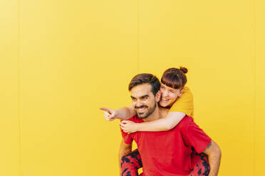 Lächelnder Mann, der eine Frau vor einer gelben Wand huckepack nimmt - MGRF00473