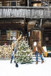Junges Paar schmückt gemeinsam den Weihnachtsbaum vor einem Bauernhaus - HHF05661