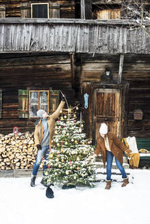 Young couple decorating Christmas tree together in front of farmhouse - HHF05661