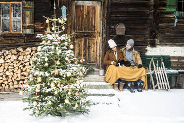 Young couple sitting with cat in front of farmhouse during winter - HHF05664