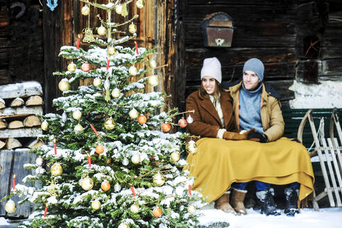 Lächelndes Paar, das vor einem Bauernhaus sitzt und den Weihnachtsbaum betrachtet - HHF05668