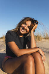 Junge Frau mit Hand im Haar am Strand sitzend - JRVF01782