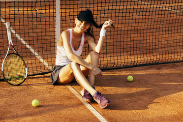 Lächelnde Tennisspielerin mit Schläger und Bällen vor dem Netz sitzend - OYF00517