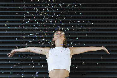 Happy businesswoman with arms outstretched throwing confetti in front of black wall - PNAF02333