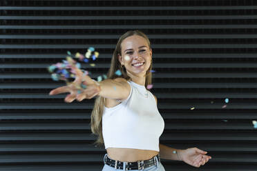 Happy female freelancer throwing confetti while standing in front of wall - PNAF02334