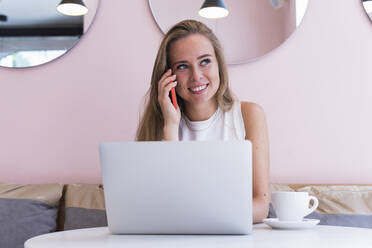 Lächelnde Freiberuflerin, die mit ihrem Laptop in einem Café sitzt und mit ihrem Handy telefoniert - PNAF02343