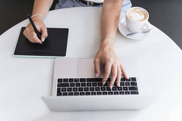 Female design professional sitting with laptop and graphic tablet in cafe - PNAF02350