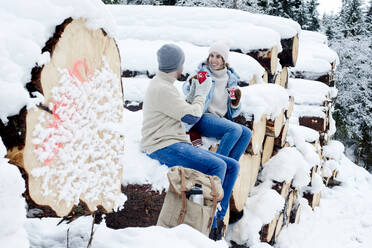 Couple enjoying coffee while sitting on snow covered logs in forest - HHF05677