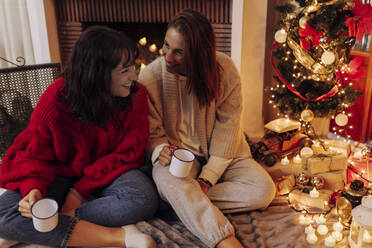 Lächelnde Freundinnen mit Tasse unter dem Weihnachtsbaum zu Hause - EGHF00174