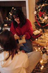 Smiling woman holding gifts while sitting with female friend at home during Christmas - EGHF00181