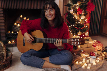 Junge Frau spielt Gitarre, während sie in der Weihnachtszeit zu Hause sitzt - EGHF00188