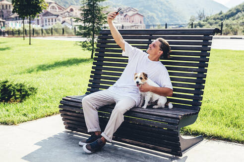 Junger Mann nimmt Selfie mit Hund durch Smartphone beim Sitzen auf Bank - OMIF00034