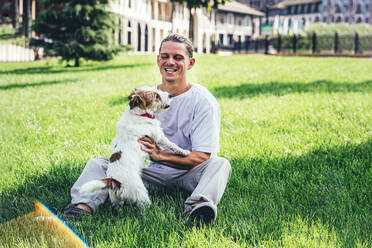 Fröhlicher Mann, der mit seinem Haustier spielt, während er auf dem Rasen sitzt - OMIF00037
