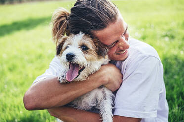Fröhlicher Mann mit geschlossenen Augen, der einen Hund auf einer Wiese umarmt - OMIF00044