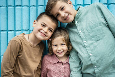 Lächelnde Geschwister, die sich aneinander lehnen, während sie vor einer blauen Wand stehen - RCPF01265