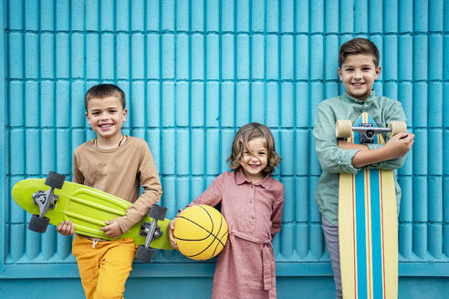 Boys and girl holding skateboards and sports ball in front of blue wall - RCPF01269